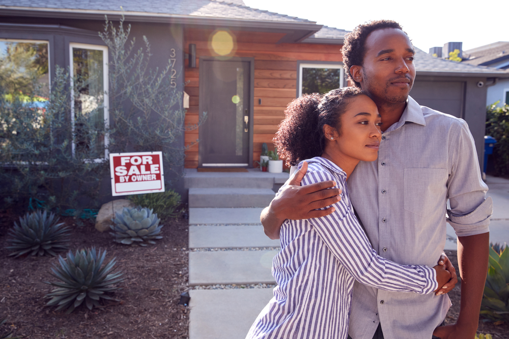 Young couple looking in the distance standing outside home with for sale sign in yard