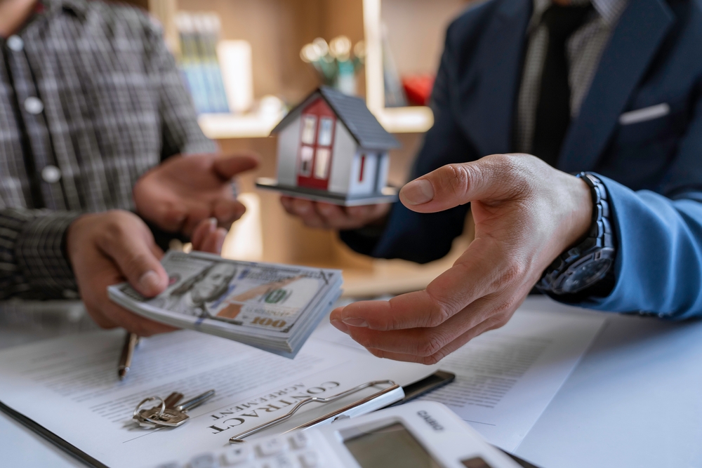 Two sets of hands, one with cash, one with house. Contract and house keys on table below.